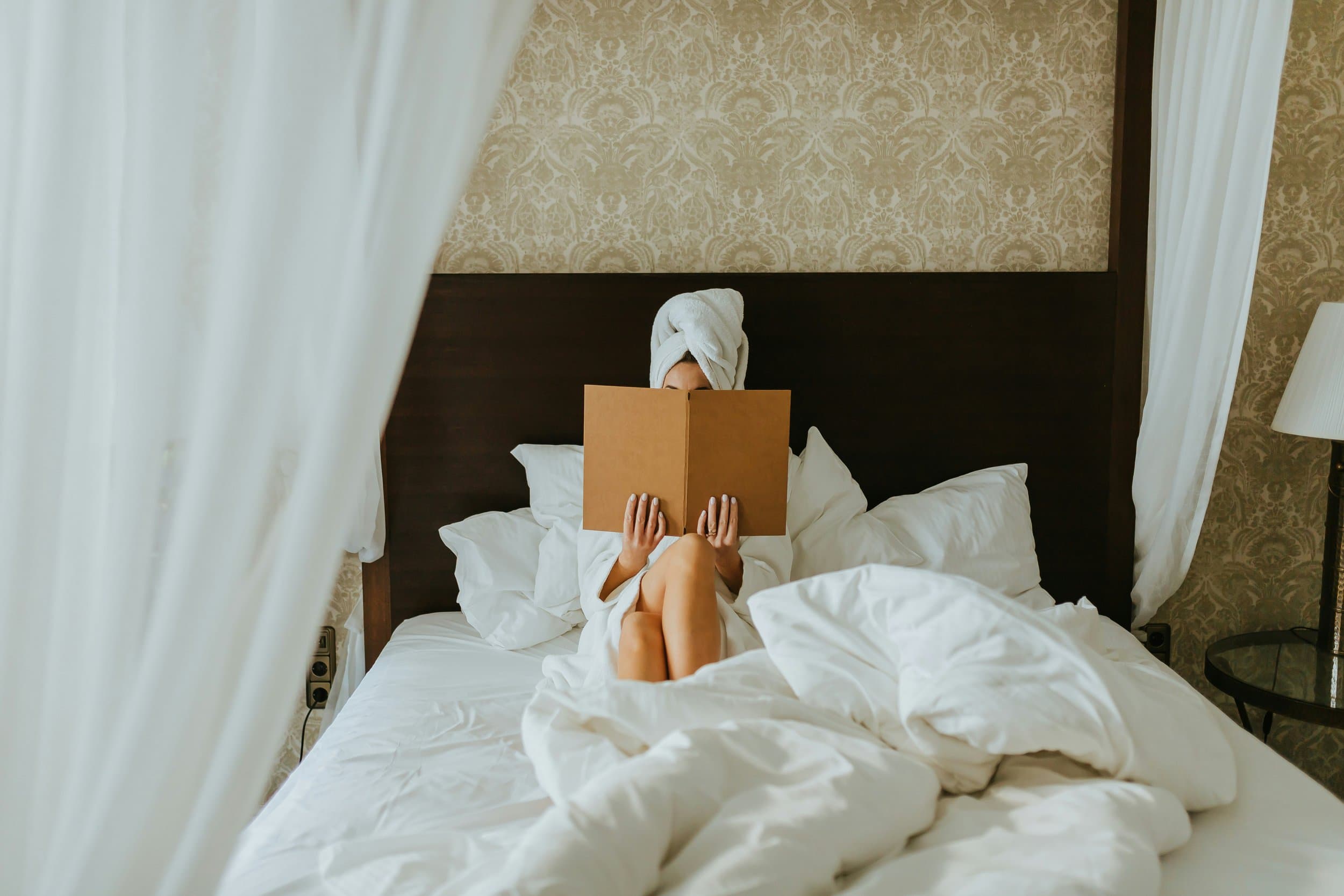 Person sitting on a bed with a towel wrapped around their head, reading a book in a hotel room.