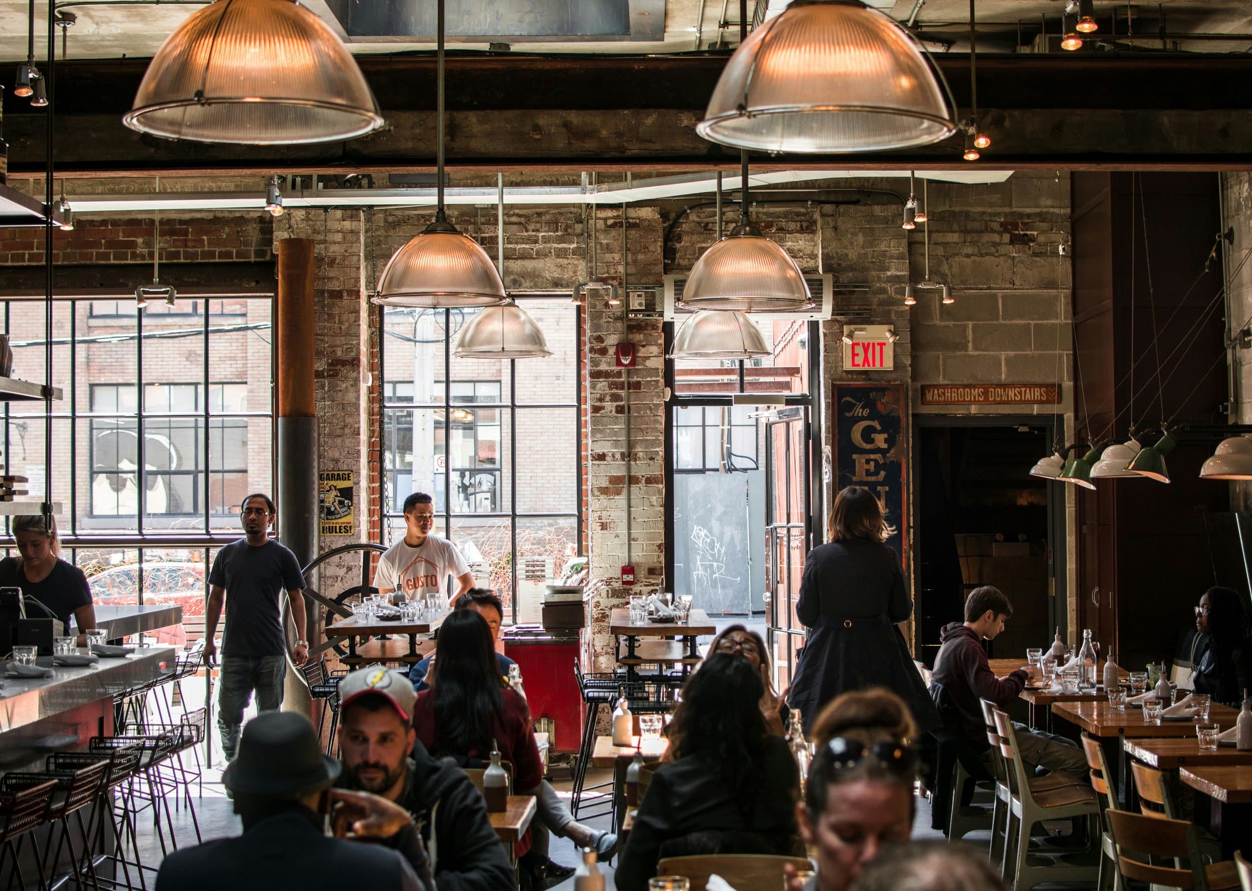 Inside a bustling restaurant with exposed brick walls, large industrial light fixtures, and large windows letting in natural light.